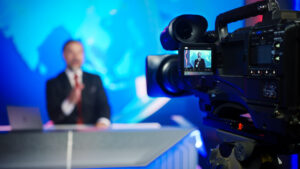 Moral Panic 22 Professional TV Camera Standing in Live News Studio with Anchor seen in Small Display. Unfocused TV Broadcasting Channel with Presenter, Newscaster Talking. Mock-up Television Channel Newsroom Set