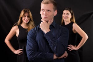 11 Warning Signs He's Actually a Player 4 A confident man doing a thinking pose and smirking while there are two smiling women looking at him in the background.