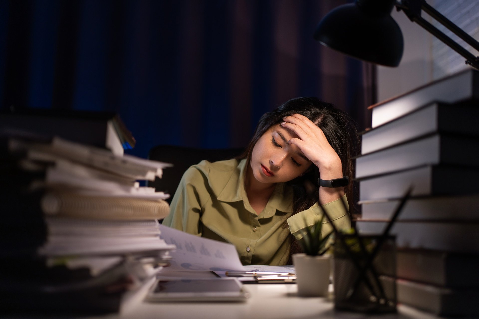 A stressed woman sat at a desk surrounded by papers and books, working late at night.